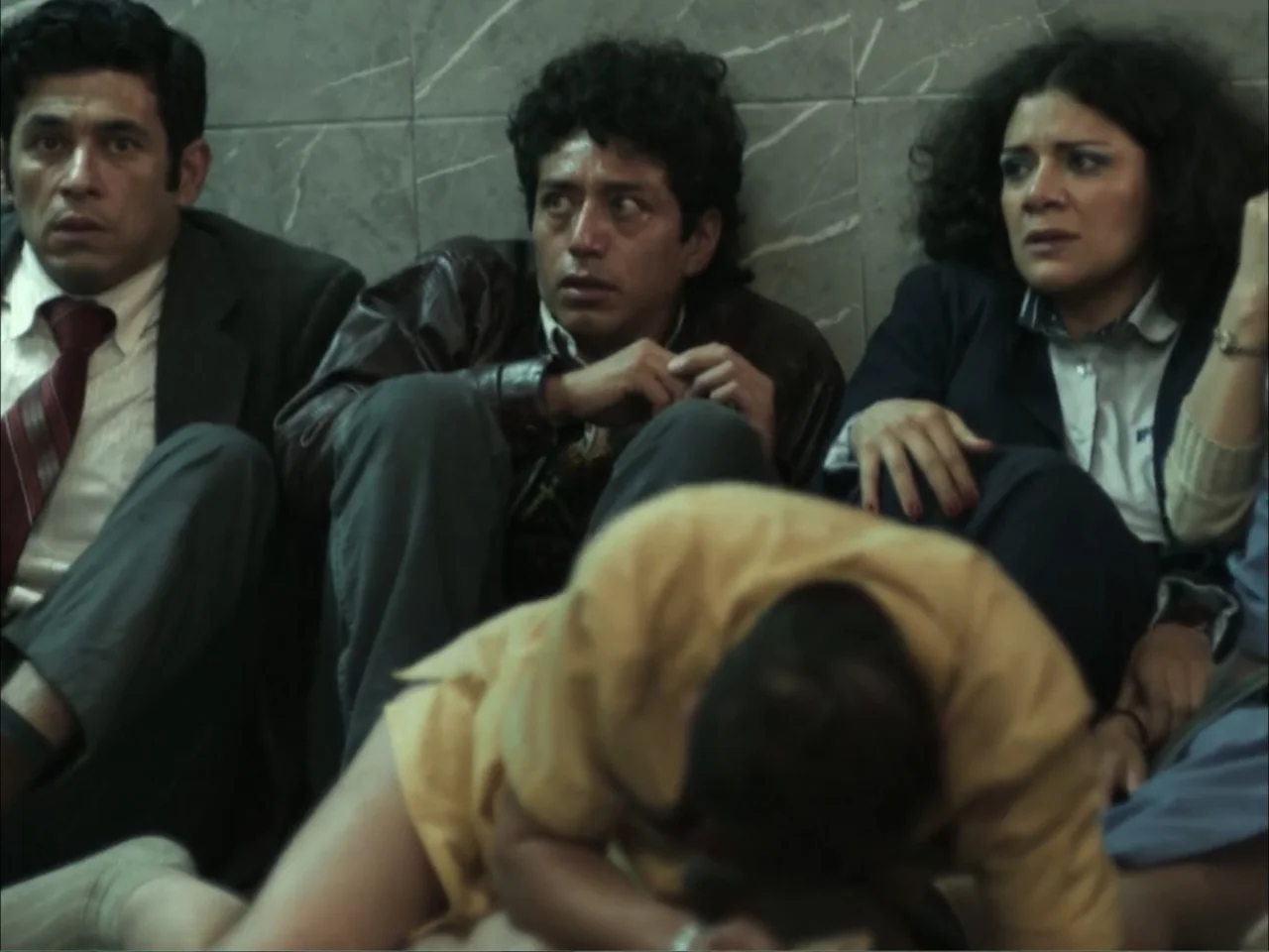 November (2025) movie still: medium — A group of people sit on the floor against a wall with marbled tiles. They are huddled together in a…