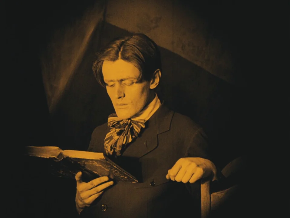 The Cabinet of Dr. Caligari (1920) movie still: medium — A man sits in a dimly lit, dark-walled room holding an open book. He is wearing a formal…
