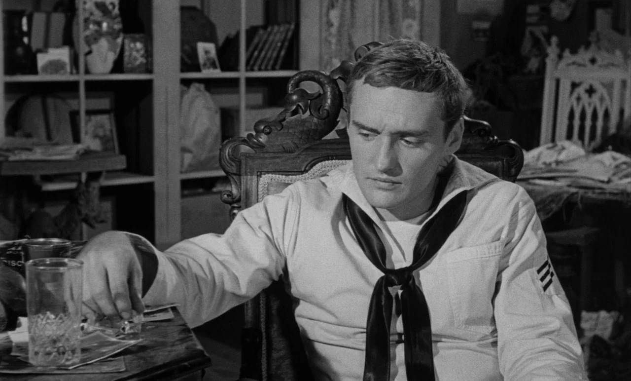Night Tide (1961) movie still: medium — A cluttered interior space features shelves filled with books and personal items. A young sailor in uniform…