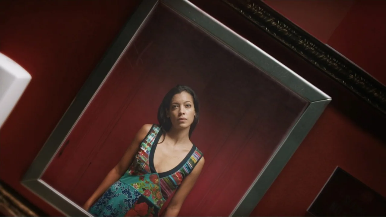 Alice in Marialand (2014) movie still: medium — A woman stands within a large rectangular picture frame hanging on a deep red wall. She gazes…