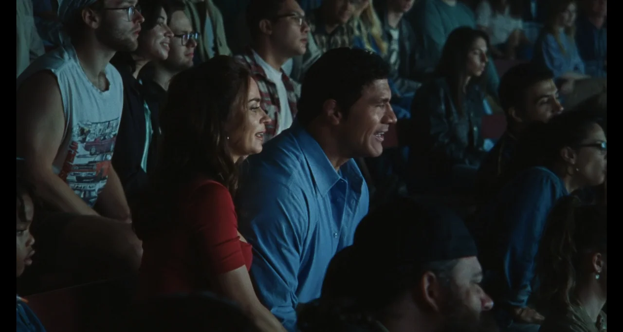 The Smashing Machine (2025) movie still: medium — A man and woman sit together on crowded stadium bleachers in low-light conditions. They both look…