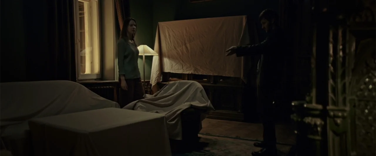 La herencia (2010) movie still: wide — A dark living room filled with furniture covered by white sheets. A man in a dark jacket gestures towards a…