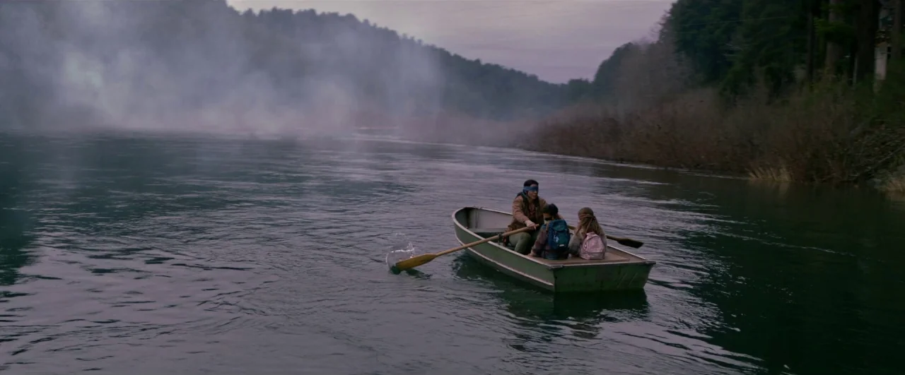 Bird Box (2018) movie still: wide — A calm, wide river flows between dense, misty hills beneath a gray sky. Three people in a small rowboat travel…
