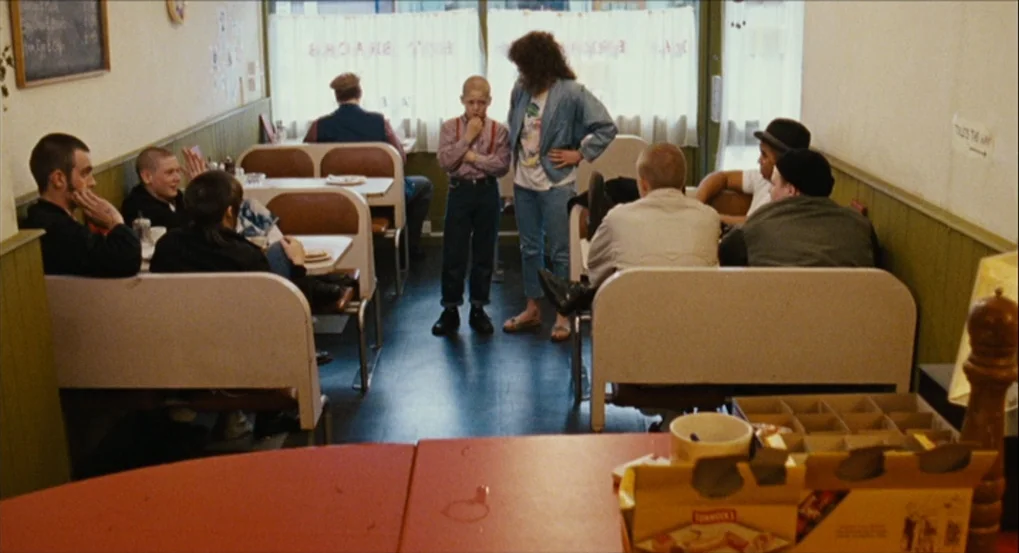 This Is England (2006) movie still: wide — A cafe with booths, pale green walls, and large windows with thin white curtains. A young boy in…