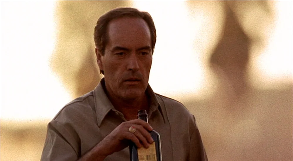 U-Turn (1997) movie still: medium — An open outdoor field is illuminated by bright daylight with a blurred background. A middle-aged man in a tan…