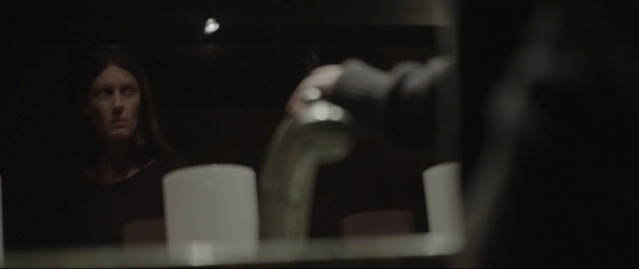 Sun Choke (2015) movie still: medium — A dimly lit room with a reflective surface showing a woman looking forward. A hand reaches toward a faucet to…
