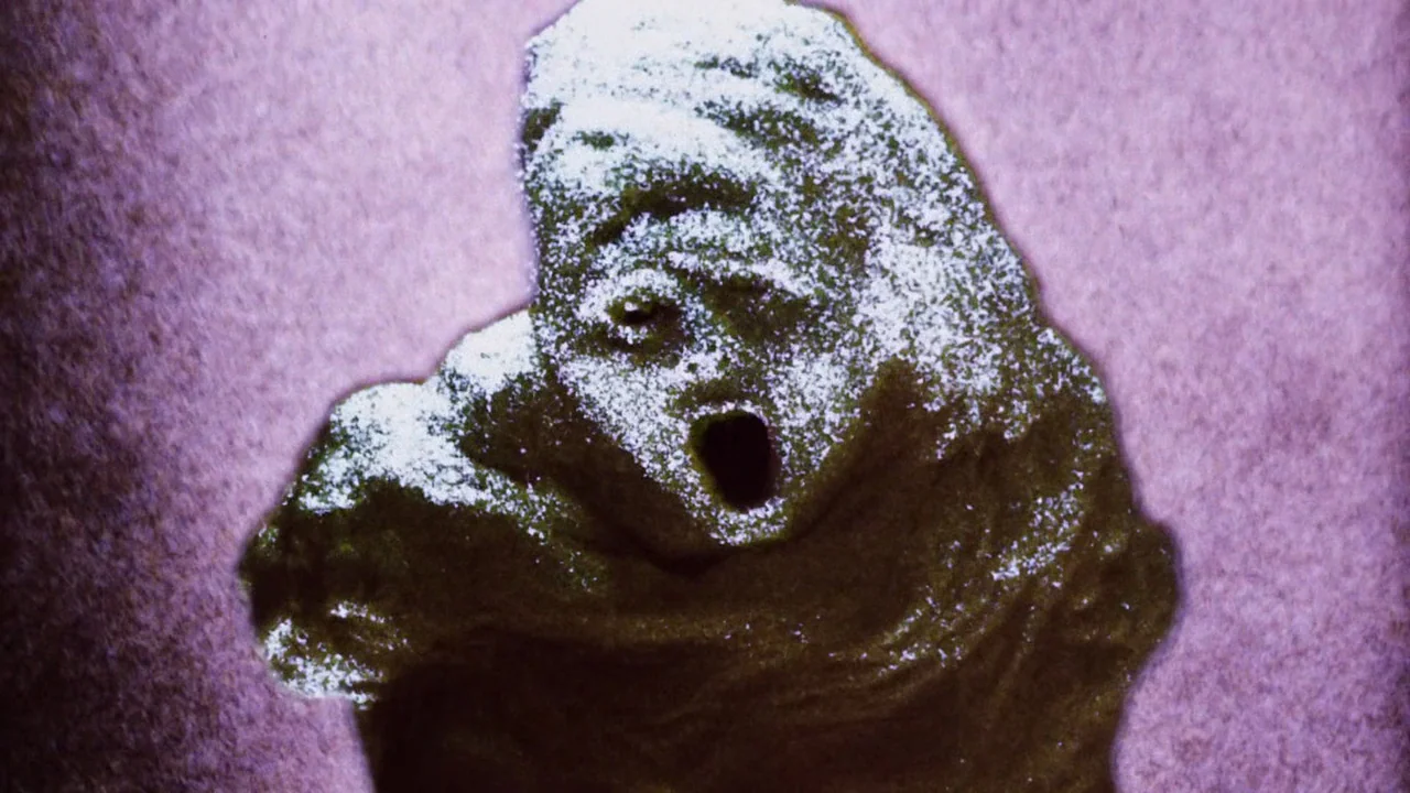 Altered States (1980) movie still: closeup — A dark, uneven, dough-like material forms the shape of a screaming humanoid head. The object rests…