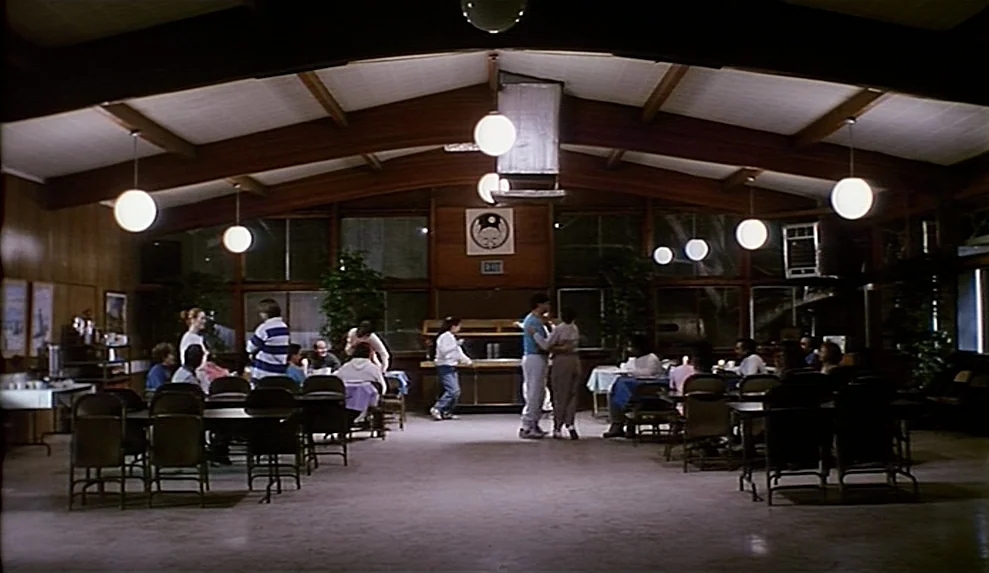 Safe movie still: wide — A spacious community hall features wood-paneled walls and exposed ceiling beams with spherical lights. Several people…