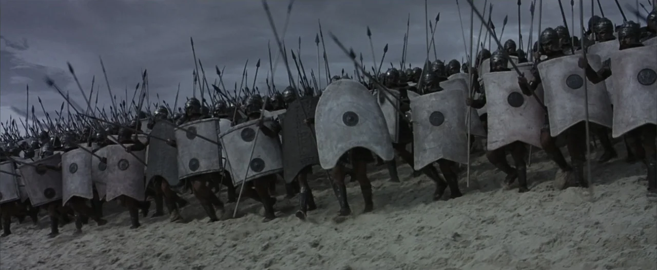Troy (2004) movie still: wide — A dense formation of ancient soldiers marches across a bleak sandy beach under clouds. The infantry carries tall…