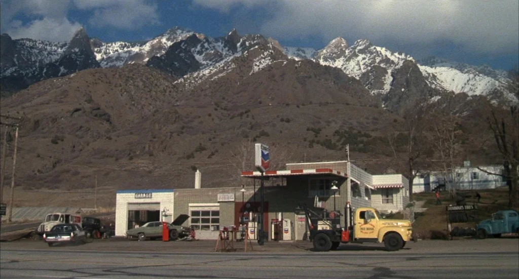 Melvin & Howard (1980) movie still: wide — A rural gas station sits at the base of large, snow-capped rocky mountains. Several vintage cars and a…