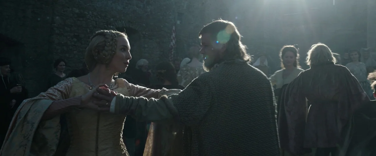 The Last Duel (2021) movie still: medium — A crowded stone castle courtyard with many guests standing in the blurred background. A noble woman and a…