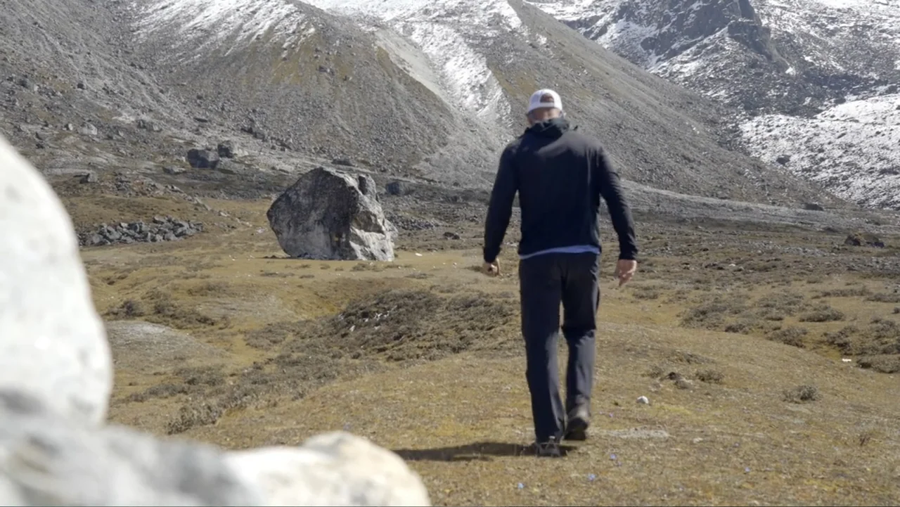 Drop Zone Everest (2023) movie still: fullbody — A vast, rocky alpine valley lies beneath towering, snow-dusted mountain slopes. A lone hiker walks…