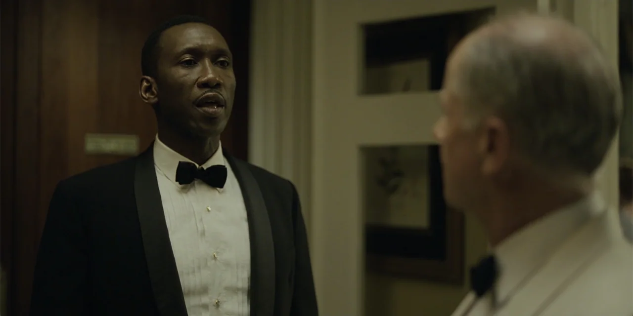 Green Book (2018) movie still: medium — An interior hallway features dark wood paneling and recessed wall nooks with framed artwork. A black man in…