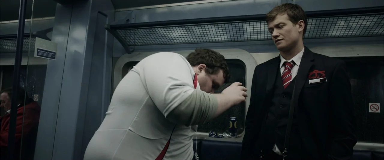 Howl (2015) movie still: medium — An interior train carriage with blue plastic seating and metal overhead luggage racks. A man in a formal suit…