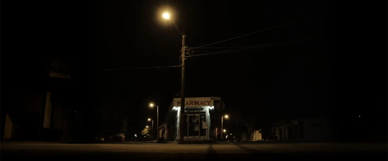 At Dawn (2013) movie still: wide — A quiet street intersection at night is illuminated by tall utility pole streetlamps. A small pharmacy building…