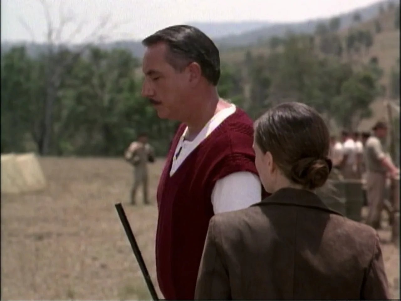 In Pursuit of Honor (1995) movie still: medium — A dry, grassy military field with tents and distant figures in the background. A man in a maroon…