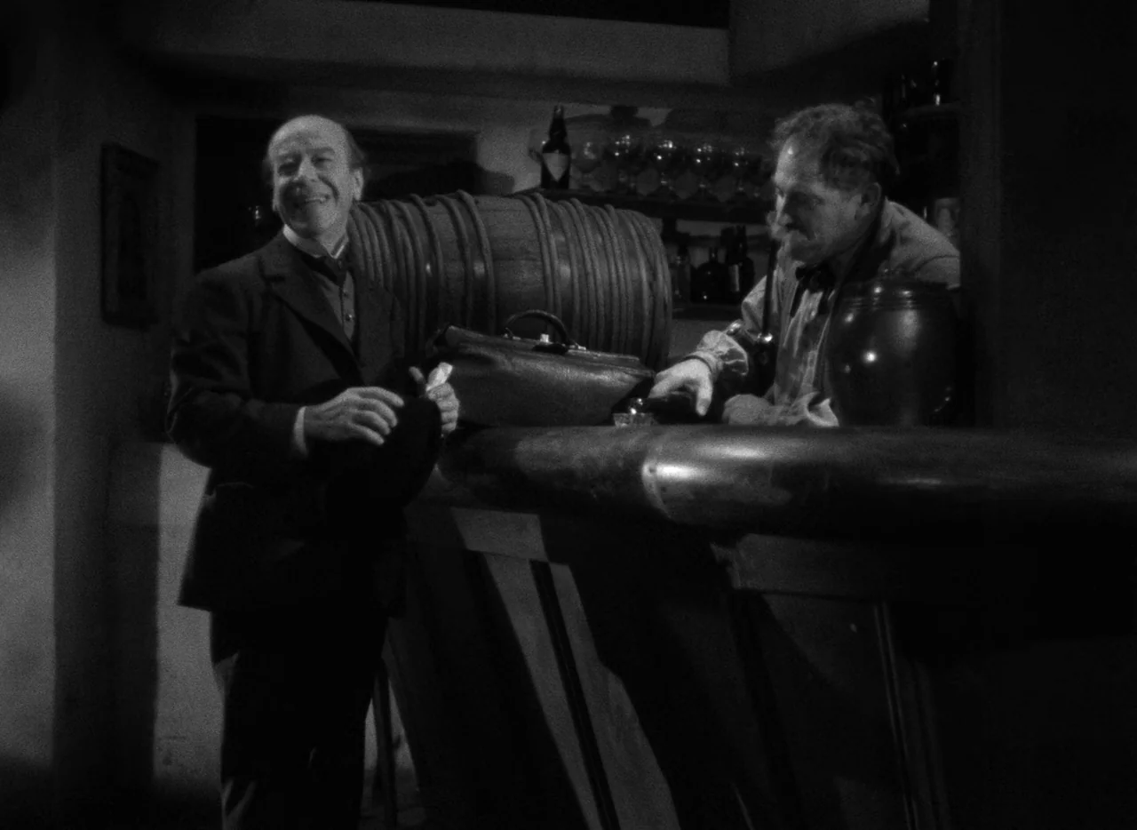 Mark of the Vampire (1935) movie still: medium — A dimly lit room with a wooden counter and a large barrel behind it. A man in a suit stands before…