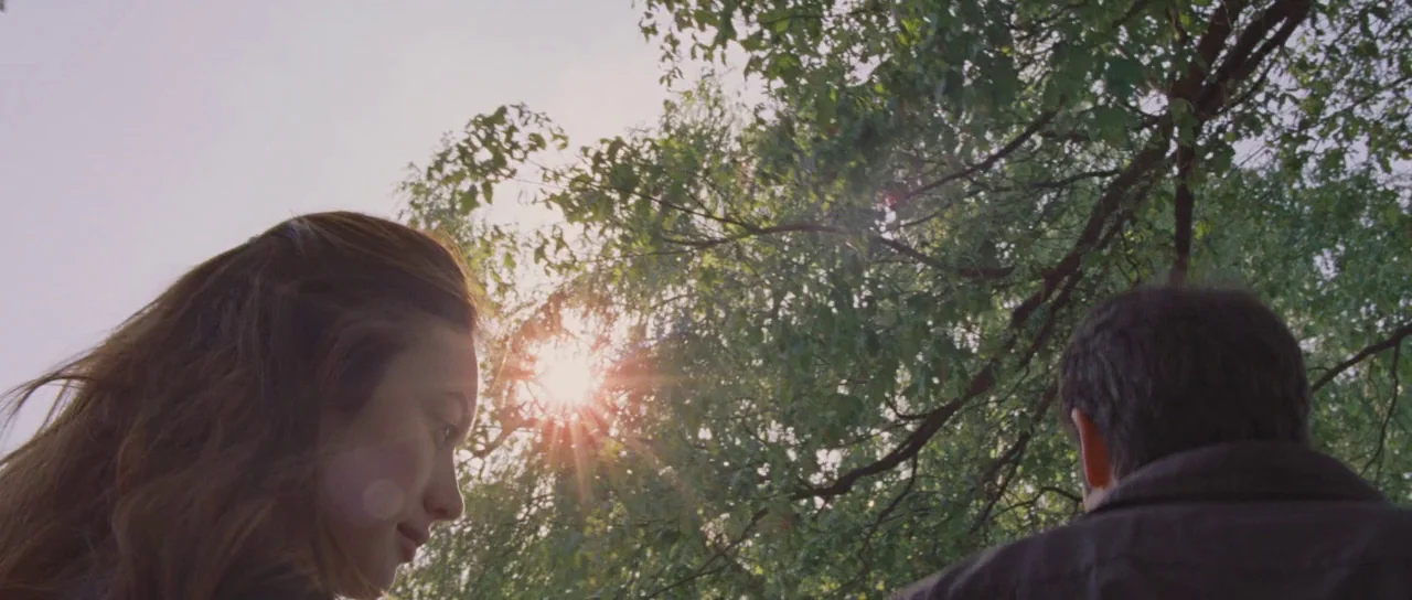 To The Wonder (2012) movie still: medium — A man and woman stand beneath a dense green canopy of tree branches outdoors. Sunlight flares intensely…