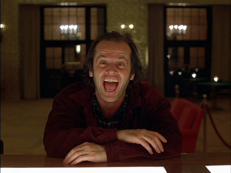 The Shining (1980) movie still: medium — A dimly lit hotel bar features dark wood accents and red cushioned chairs. A middle-aged man leans across…