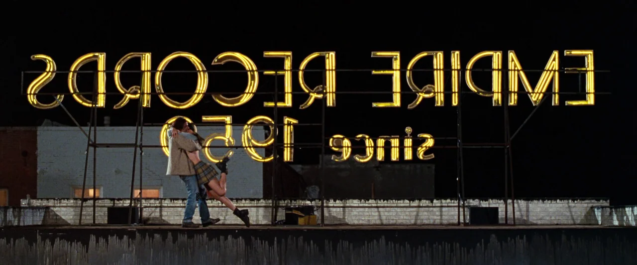 Empire Records (1995) movie still: fullbody — A tar-covered flat roof features a large, back-lit neon sign for a record shop. A man and woman…