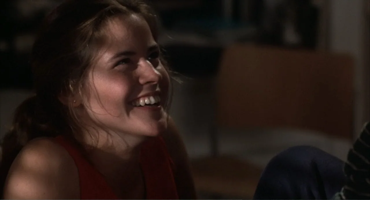 WarGames (1983) movie still: closeup — A dimly lit indoor space with blurred furniture pieces visible in the background. A young woman wearing a red…