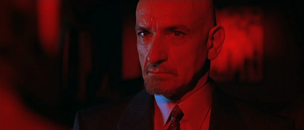 Sexy Beast (2000) movie still: closeup — A man in a suit stands in a dark environment with red lighting. He stares forward while bathed in intense…