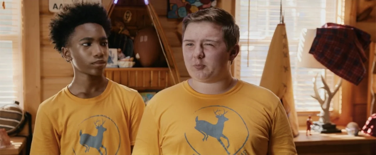 Camp Hideout (2023) movie still: medium — A rustic interior with log walls and wooden surfaces holds a variety of camp props. Two teenage boys…