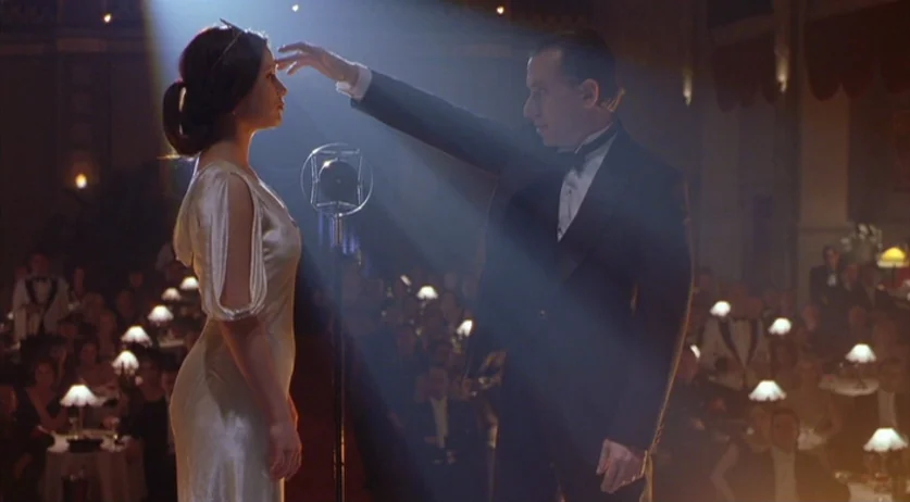 Invincible (2001) movie still: medium — A crowded dimly lit nightclub room with small lamps on tables. A man in a tuxedo stands performing a…