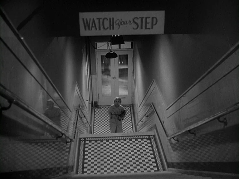Killer’s Kiss (1955) movie still: wide — A tiled indoor stairwell with a prominent sign reading 'WATCH YOUR STEP' above. A woman in a light trench…