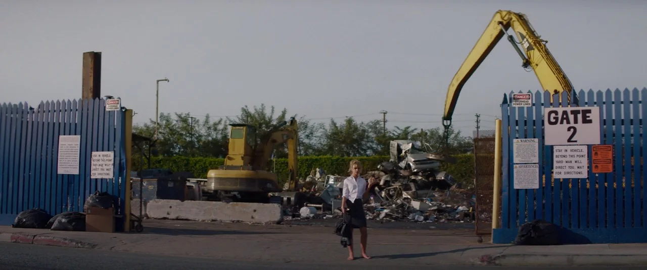 Promising Young Woman (2020) movie still: wide — A fenced-in junkyard with piles of scrap metal and large yellow excavators. A lone woman stands at…