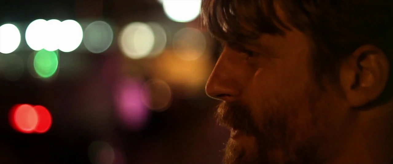 Bellflower (2011) movie still: closeup — The man stands on a city street at night near blurred city lights. He faces right while his features are…