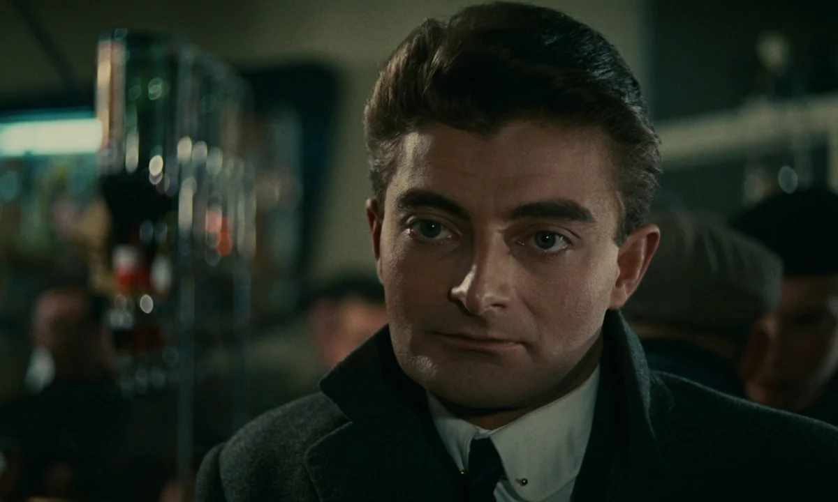 Muriel (1963) movie still: closeup — A dimly lit bar with blurred bottles visible on shelves in the background. A man stands facing forward against…