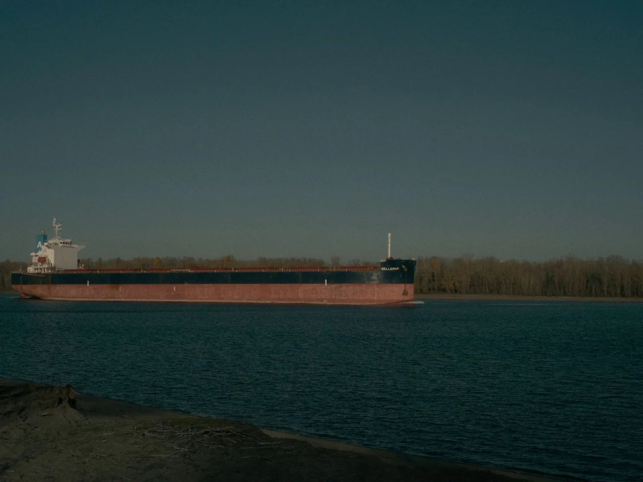 First Cow (2019) movie still: wide — A long freighter travels down a wide river lined with bare autumn trees. The vessel features a dark hull and a…