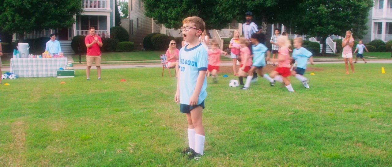 Greener Grass (2019) movie still: fullbody — A suburban residential neighborhood with large houses visible behind a green grass field. A young boy…