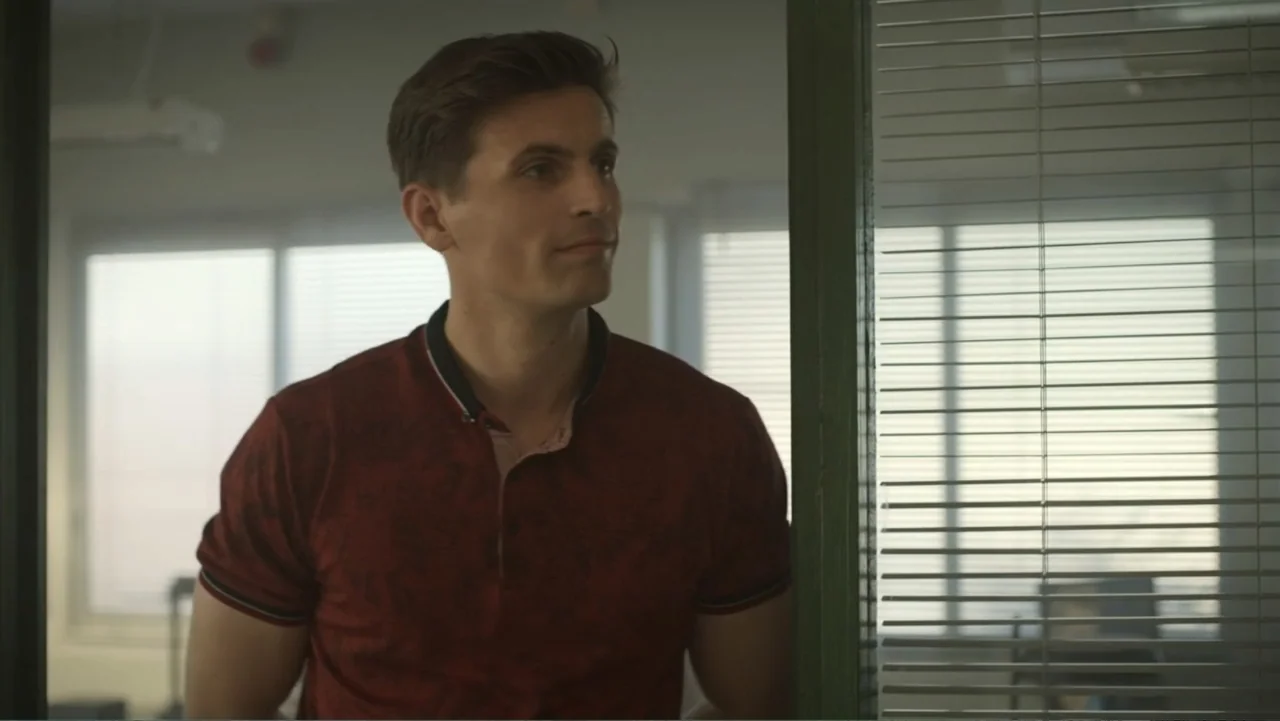 Unknown #1 movie still: medium — An office interior features a glass door frame and windows covered with horizontal blinds. A young man in a dark…
