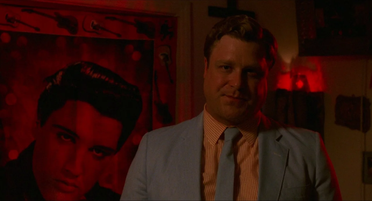 True Stories (1986) movie still: medium — A man stands before a large, prominent poster of Elvis Presley with guitars. The room is bathed in an…