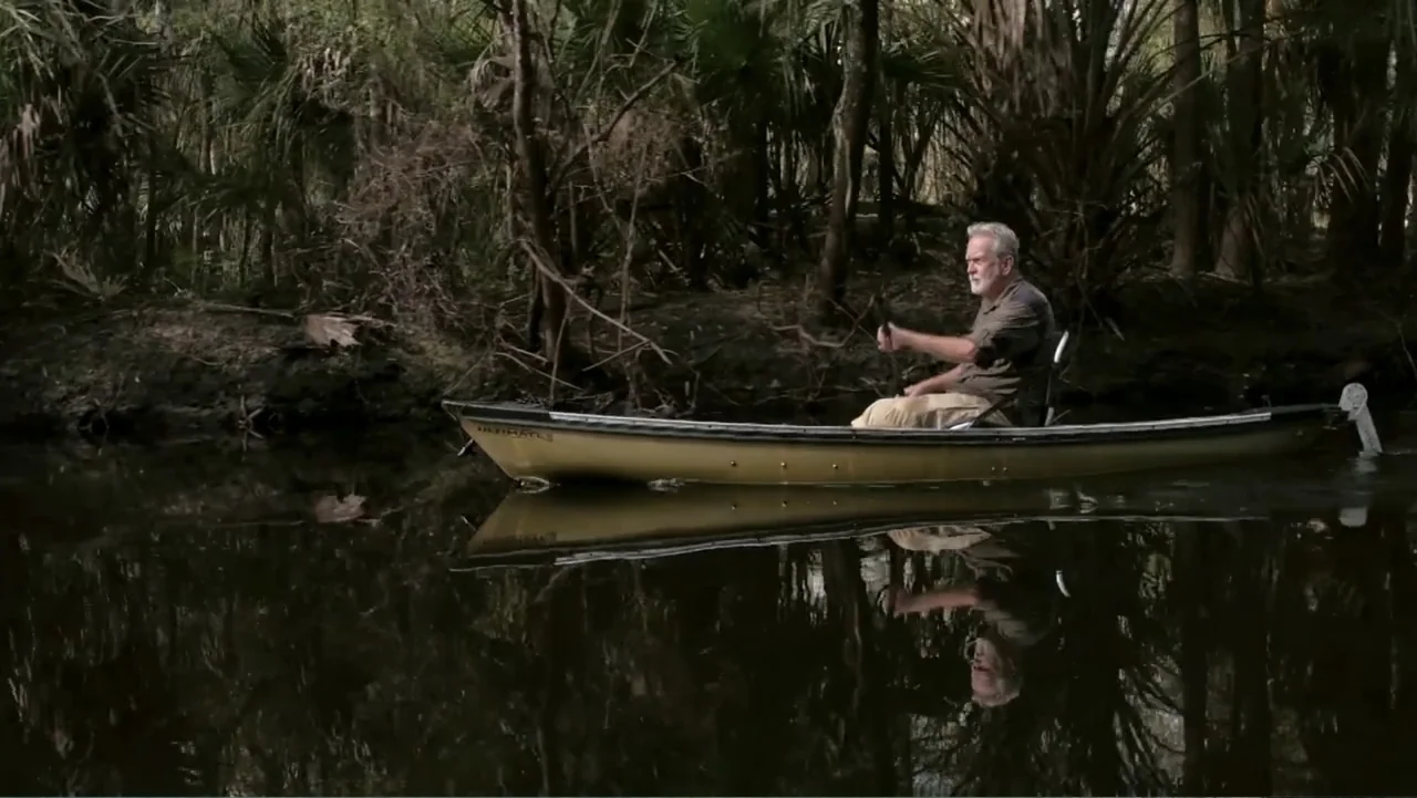 The Forever Prisoner (2021) movie still: medium — A man paddles a tan kayak through a calm swamp filled with dark vegetation. The subject sits in…