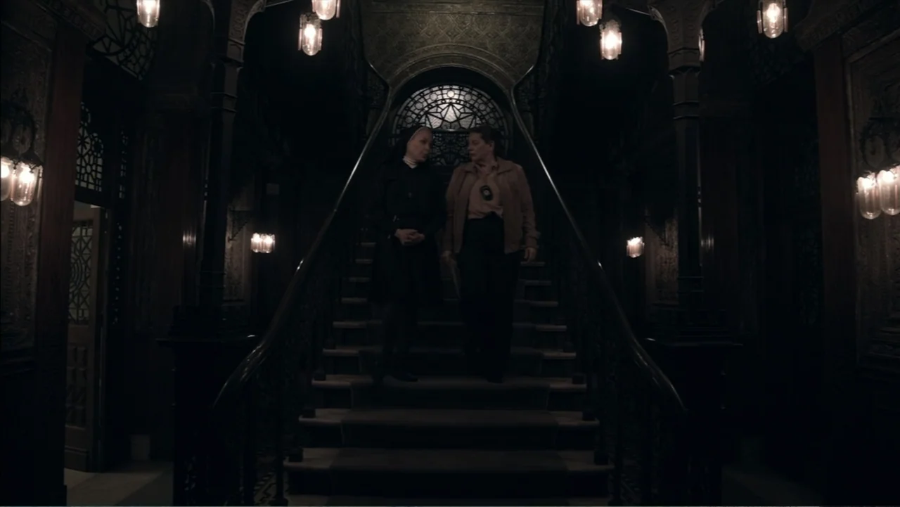 The Grotto (2022) movie still: fullbody — An ornate dark interior features a central grand staircase with decorative wooden banisters. A nun and a…