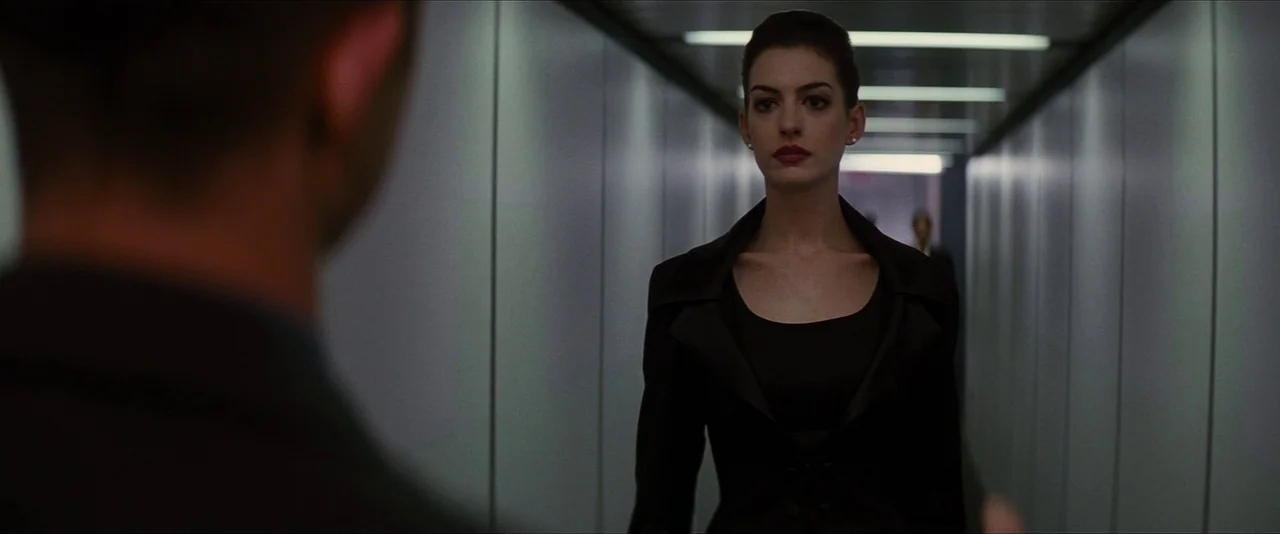 The Dark Knight Rises (2012) movie still: medium — A narrow airport jet bridge is lined with flat metal panels and bright ceiling lights. A woman in…