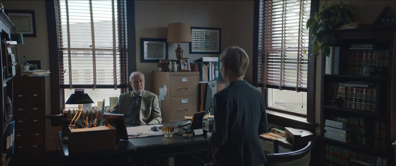 Rebel Ridge (2024) movie still: medium — A cluttered law office with rows of books and venetian blinds over the windows. An elderly man sits behind…