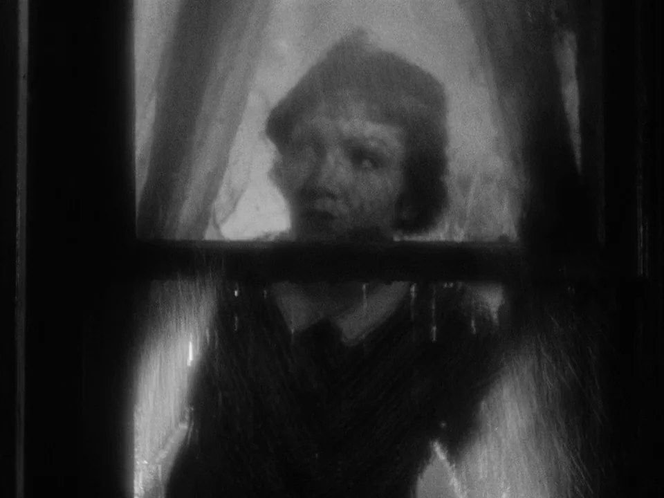 It Happened One Night (1934) movie still: medium — A woman looks through a rain-streaked window pane from inside a dark room. The view is obscured…