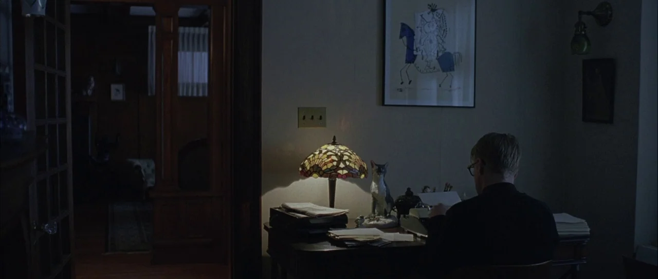 Capote (2005) movie still: medium — A dark home office is lit primarily by a stained glass lamp on a desk. A man with blonde hair sits at the desk…