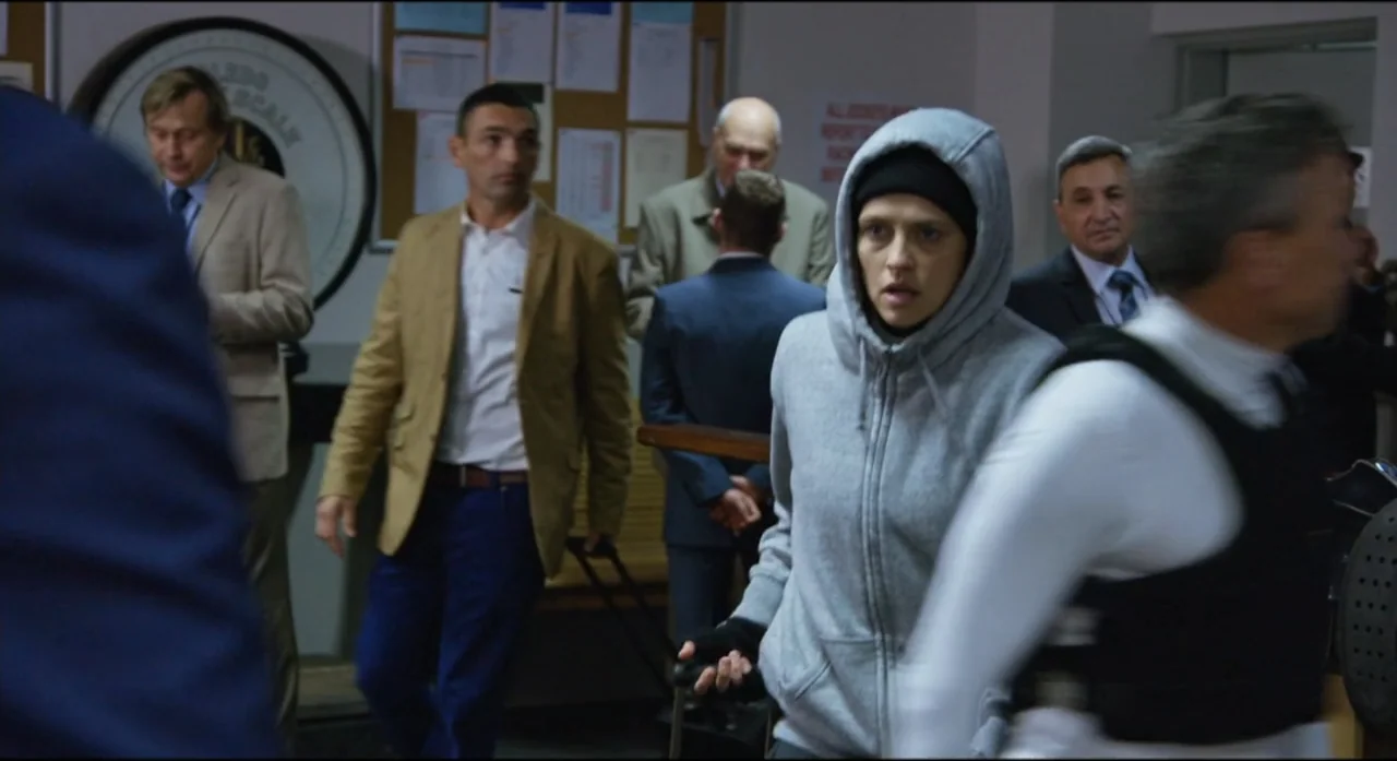 Ride Like a Girl (2019) movie still: medium — A busy government lobby with beige walls, a circular plaque, and several men walking. A woman in a…