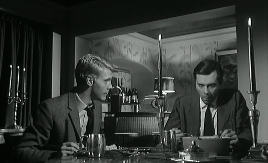 The Servant (1963) movie still: medium — A dark dining room with patterned wallpaper and a bar area in the back. Two men in suits sit at a round…