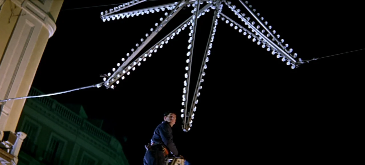 Live Flesh (1997) movie still: wide — A night view of a man positioned on a lift near a large star-shaped decoration. The bright neon light…