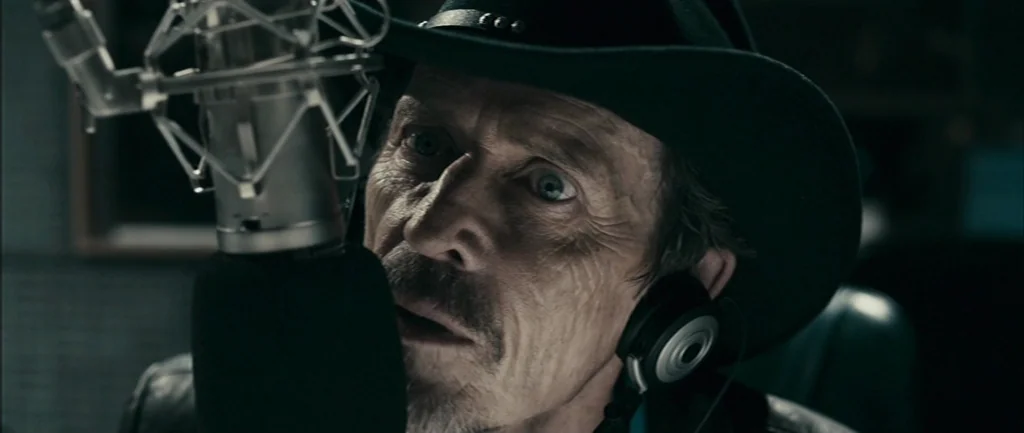 Pontypool (2008) movie still: closeup — A dimly lit recording booth with acoustic foam padding visible on the wall. A man in a cowboy hat leans…