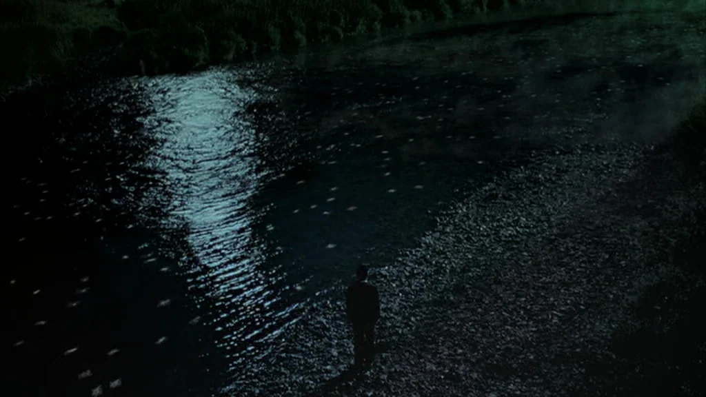 Everything Is Illuminated (2005) movie still: wide — A dark river flows past a gravel bank under moonlight. A man in a suit stands alone, facing the…