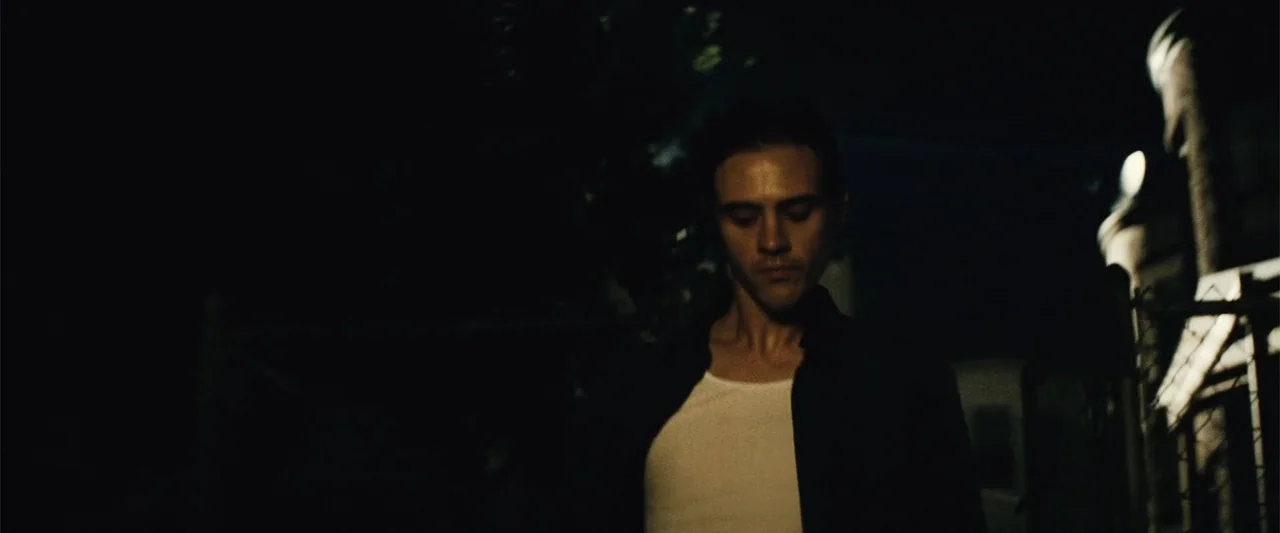 Run All Night (2015) movie still: medium — A man stands outdoors at night near a chain link fence. The man wears a white undershirt and an…