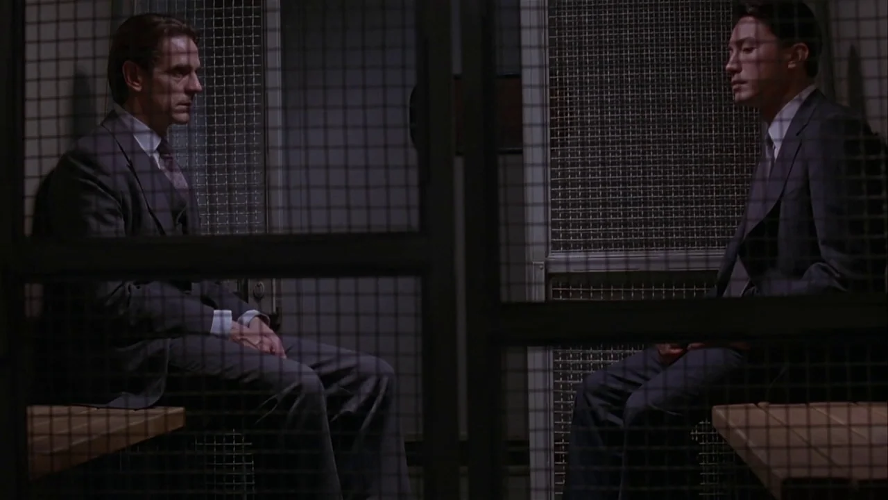 M. Butterfly (1993) movie still: medium — A visitation room features two metal-mesh screened enclosures divided by a partition wall. Two men wearing…