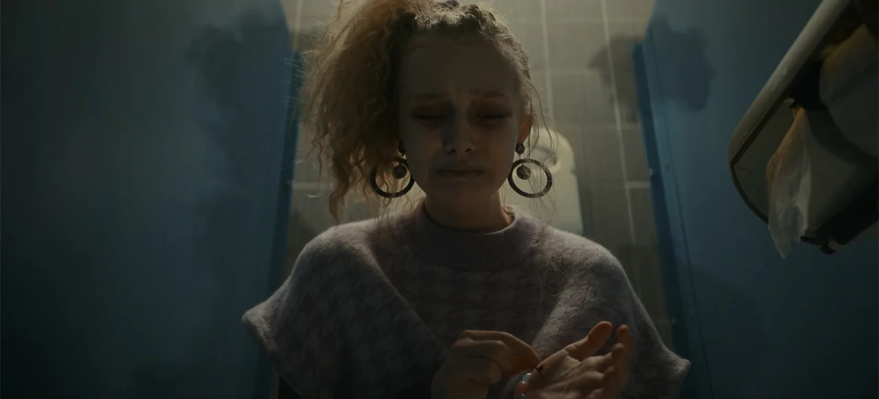 My Best Friend's Exorcism (2022) movie still: closeup — A dimly lit public restroom stall with dark blue walls and visible tile lines. A young woman…